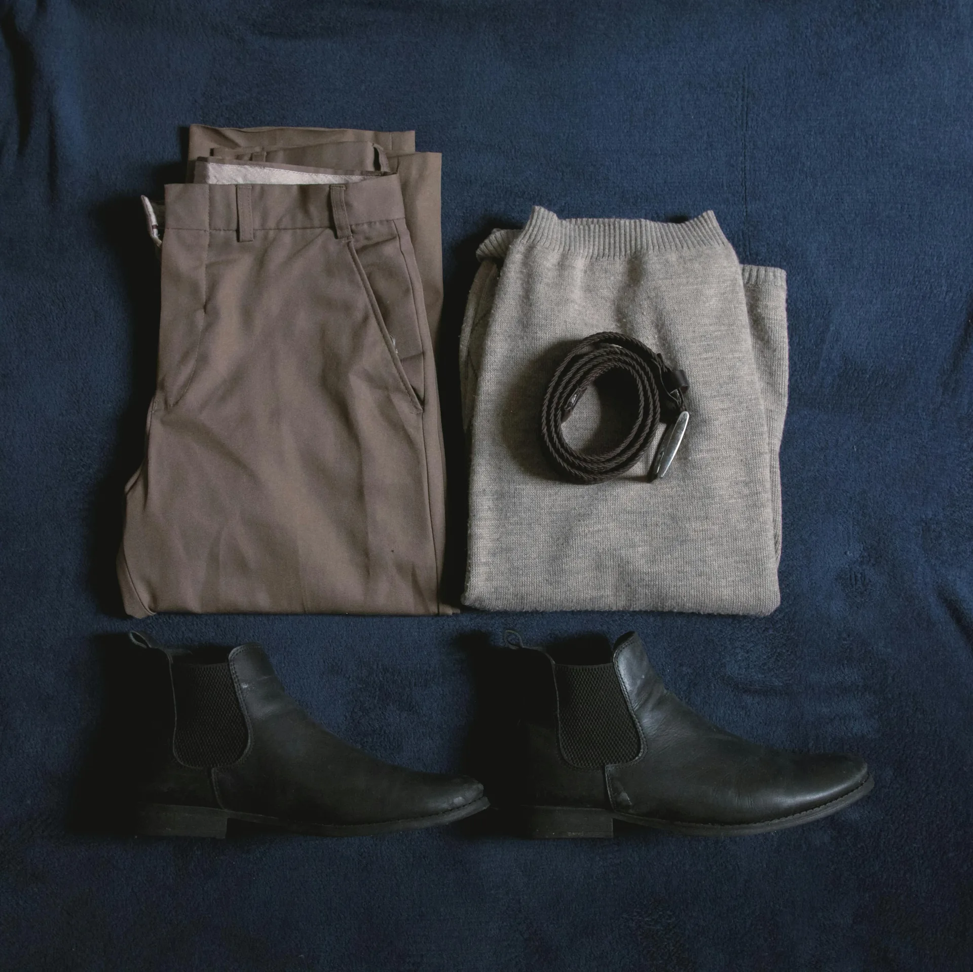 Folded neutral basics laid out: trousers, a sweater, a belt, and boots