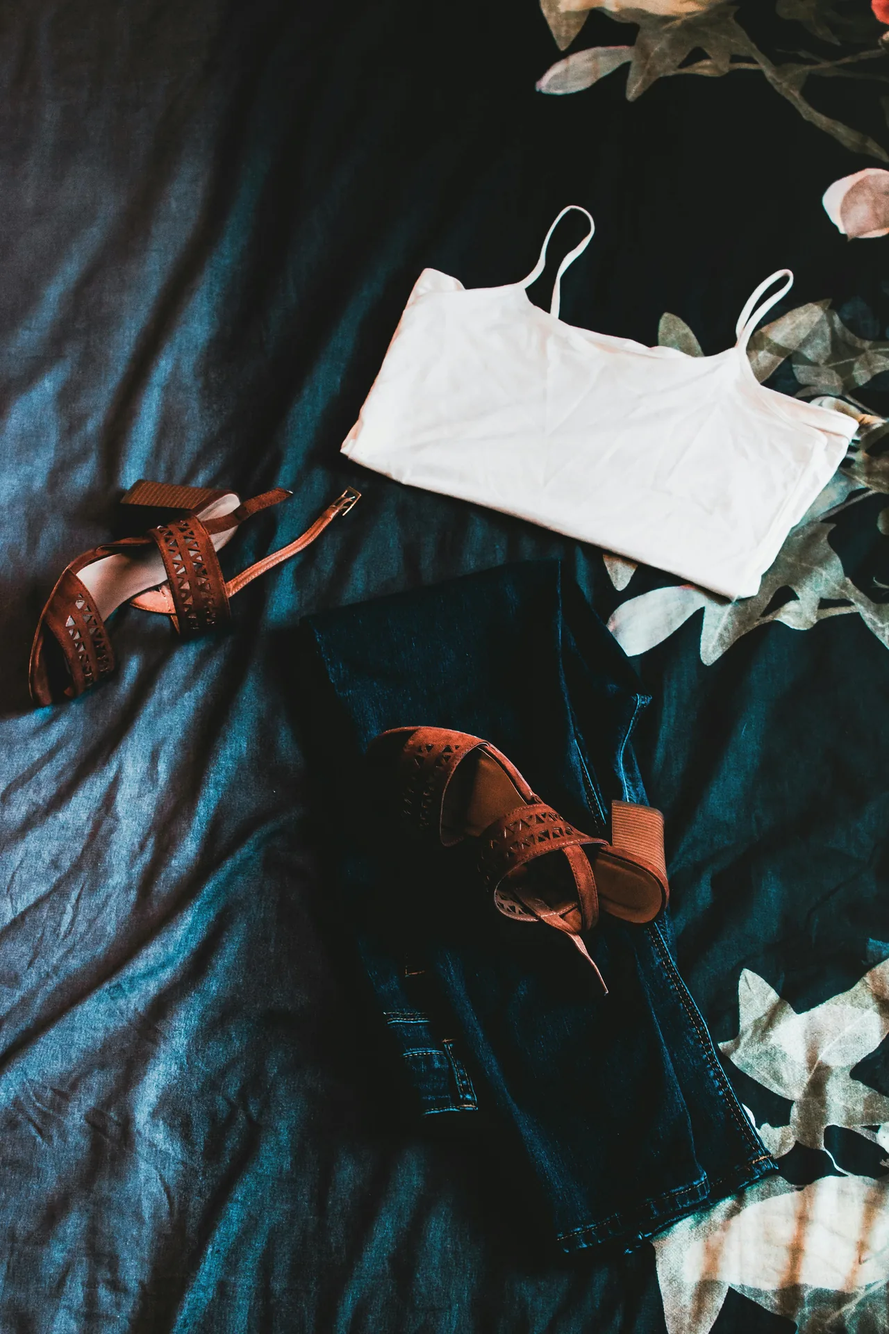 A moody outfit flat-lay: a white cami, dark jeans, and brown sandals