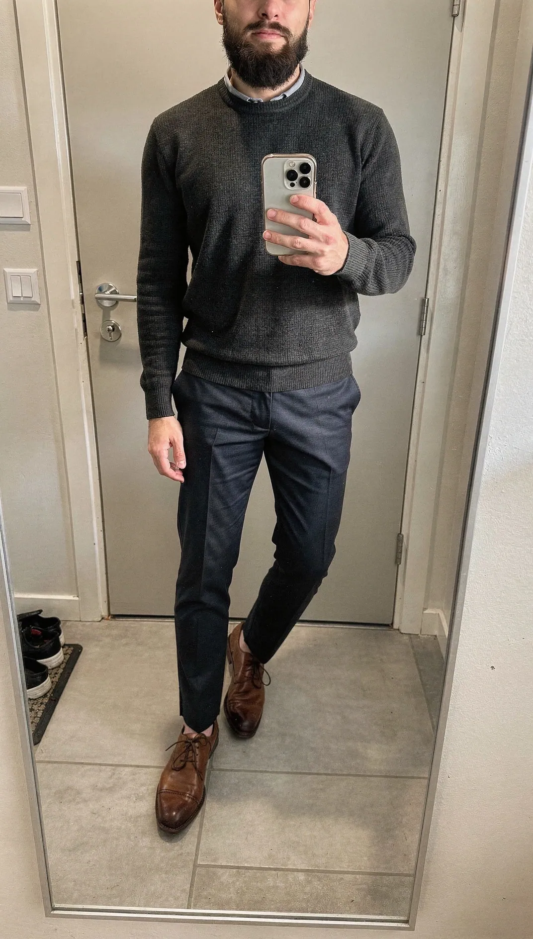A man's mirror selfie in a charcoal crew-neck sweater over a light blue collared shirt, navy trousers and brown lace-up shoes