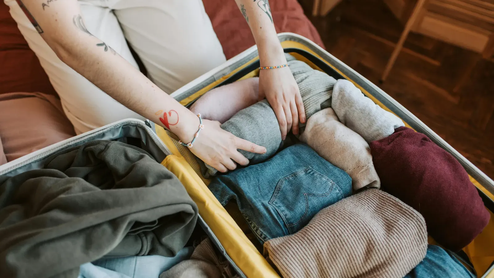 Mani che arrotolano vestiti in una valigia aperta su un letto: jeans blu e maglioni grigi, beige e bordeaux; nel coperchio, una felpa con cappuccio verde oliva
