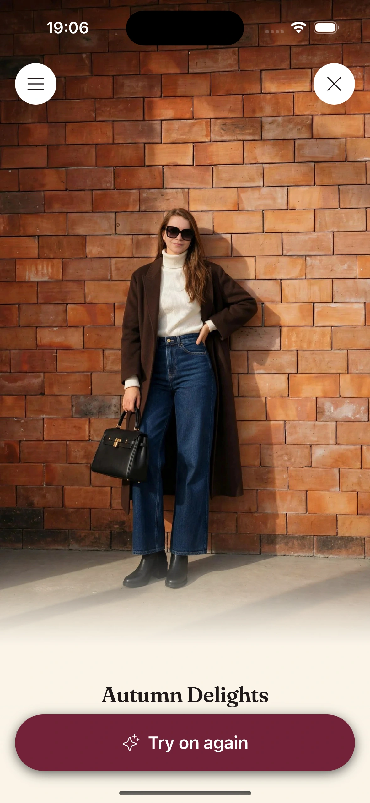 A Clottr try-on result titled Autumn Delights: a woman in a long dark brown coat over a cream turtleneck, dark wide-leg jeans, sunglasses and a black bag, leaning on an orange brick wall